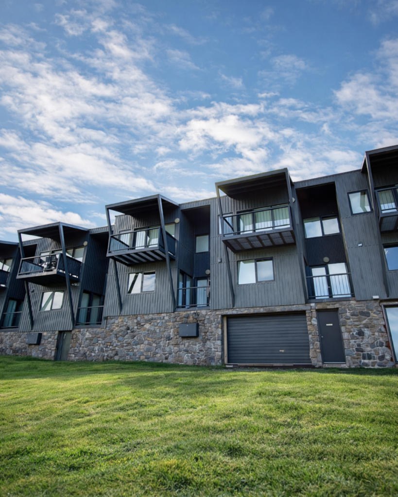Exterior of The Stables Resort in Perisher during the warmer months, featuring alpine apartments with private balconies, stone walls and green grass beneath a blue sky.
